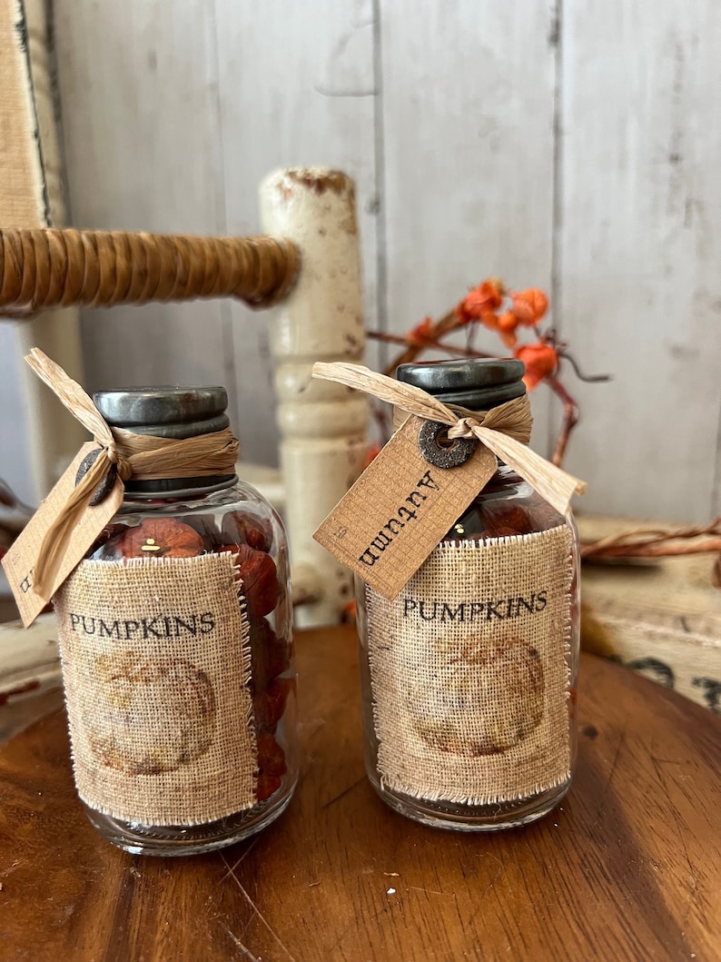 May include: Two glass jars with metal lids and burlap labels. The labels are decorated with a rustic pumpkin design and the text 'Pumpkins'. One jar has a wooden tag that says 'Autumn'. The jars are filled with small, brown objects.