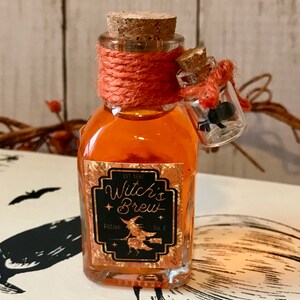 Pu&ograve; includere: Una piccola bottiglia di vetro con un tappo di sughero e liquido arancione all'interno. La bottiglia ha un'etichetta che dice "Witch's Brew Potion No. 2" con un'illustrazione di una strega. La bottiglia &egrave; legata con uno spago arancione.