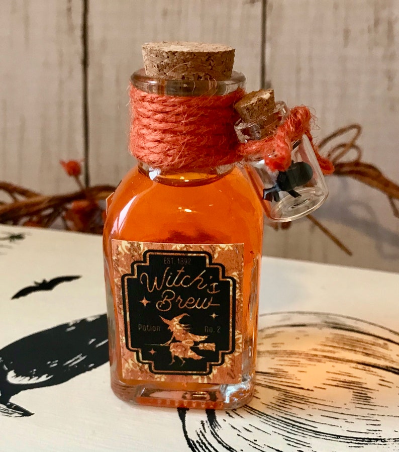 Pu&ograve; includere: Una piccola bottiglia di vetro con un tappo di sughero e liquido arancione all'interno. La bottiglia ha un'etichetta che dice "Witch's Brew Potion No. 2" con un'illustrazione di una strega. La bottiglia &egrave; legata con uno spago arancione.