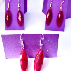 May include: Three pairs of dangle earrings with red gemstone drops. The earrings are displayed on purple card stock.