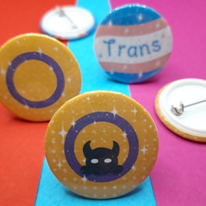 Könnte beinhalten: Vier runde Anstecknadeln mit verschiedenen Designs. Der erste Knopf ist gelb mit einem violetten Kreis und dem Text "Trans" in Blau. Der zweite Knopf ist gelb mit einem violetten Kreis und einer schwarzen Silhouette einer Kreatur mit einer Krone. Der dritte Knopf ist weiß mit einer silbernen Nadel. Der vierte Knopf ist blau mit einem weißen Streifen und dem Text "Trans" in Schwarz.