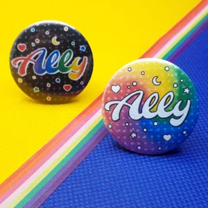 Könnte beinhalten: Zwei runde Ansteckbuttons. Der linke Button hat einen schwarzen Hintergrund mit regenbogenfarbenem Schriftzug „Ally“, kleinen Herzen, Sternen und Monden. Der rechte Button zeigt einen Regenbogenverlauf mit weißem Schriftzug „Ally“ und Symbolen.