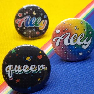 Könnte beinhalten: Drei runde Ally- und Queen-Ansteckbuttons. Ein Button hat einen Regenbogenhintergrund mit dem Wort "Ally" in weißer Schrift. Ein weiterer Button hat einen schwarzen Hintergrund mit dem Wort "Ally" in Regenbogen-Schrift. Der dritte Button hat einen schwarzen Hintergrund mit dem Wort "queen" in Regenbogen-Schrift.