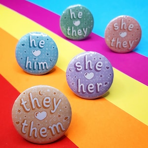 May include: Five round buttons with different pronoun combinations. The buttons are decorated with hearts, stars, and moons. The text on the buttons reads: "he him", "she her", "they them", "he they", and "she they".