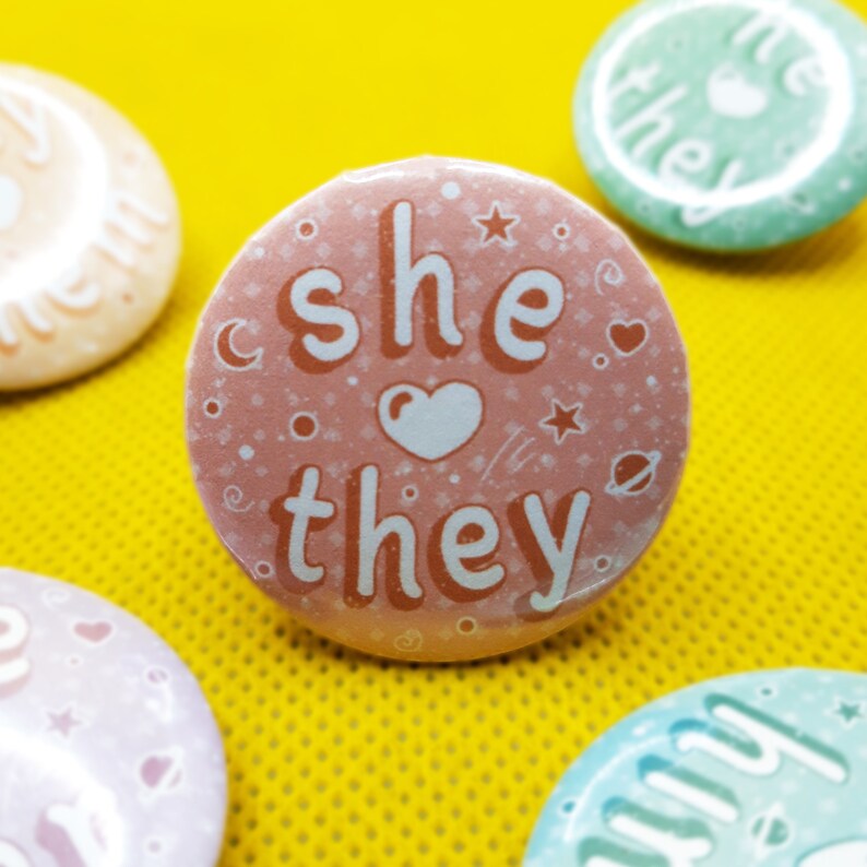May include: A pink button with a white heart and the text "she they" in orange. The button has a white background with small white stars and other symbols.