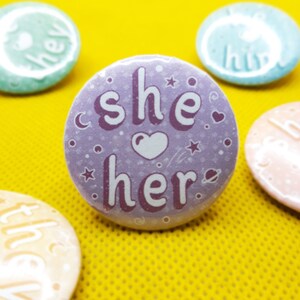 May include: Purple button with the text "she/her" in white lettering with a white heart and stars on a purple background.