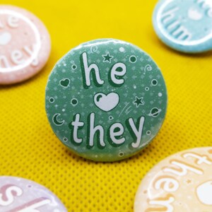 May include: Green button with white text that reads "he they" with a heart symbol between the words. The button has a sparkly background with white stars and a crescent moon.