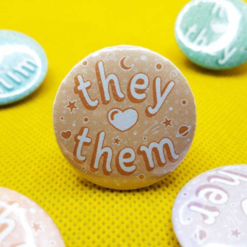 May include: A peach-colored button with the text "they them" in a white font. The button has a heart and stars around the text.