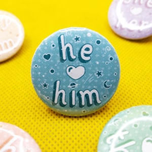 May include: A blue button with the text "he him" in white lettering with a white heart in between the words. The button has a light blue background with white stars and a crescent moon.