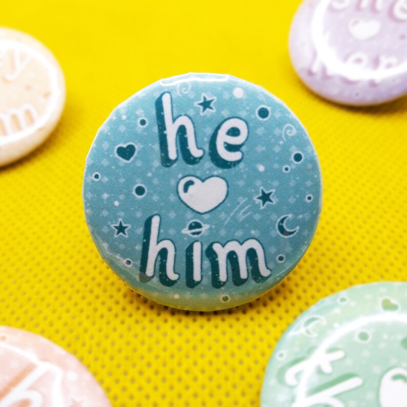 May include: A blue button with the text "he him" in white lettering with a white heart in between the words. The button has a light blue background with white stars and a crescent moon.
