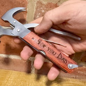 May include: A multi-tool featuring a hammer, pliers, and other implements. The wooden handle is engraved with the words "For You Dad". The tool is silver and brown, held against a brick wall.