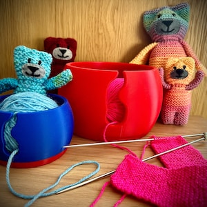 May include: A blue and red yarn bowl with blue and pink yarn. Knitted teddy bears in various colours are in the background. Knitting needles and pink knitted fabric are in the foreground.