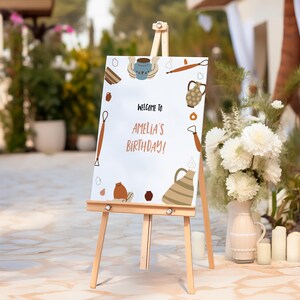 Könnte beinhalten: Eine Holzstaffelei zeigt ein weißes Schild mit dem Text "WELCOME TO AMELIA'S BIRTHDAY!" umgeben von Illustrationen zum Thema Keramik. Das Schild befindet sich im Freien, mit Blumen und Kerzen im Hintergrund.