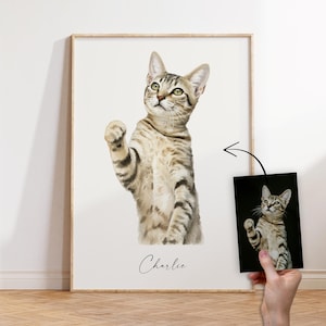 Könnte beinhalten: Aquarellporträt einer getigerten Katze mit grünen Augen, die eine Pfote hebt. Die Katze ist in einem gerahmten Druck dargestellt, mit einem kleineren Druck desselben Bildes in der Nähe. Der Name der Katze, "Charlie", ist darunter in Kursivschrift geschrieben.