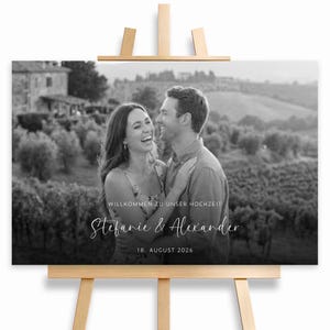 Könnte beinhalten: Schwarz-weiß Hochzeitsanzeige auf einer Holzstaffelei. Das Schild zeigt ein Foto eines lächelnden Paares mit dem Text "Willkommen zu Unser Hochzeit", "Stefanie & Alexander" und dem Datum "18. August 2026".