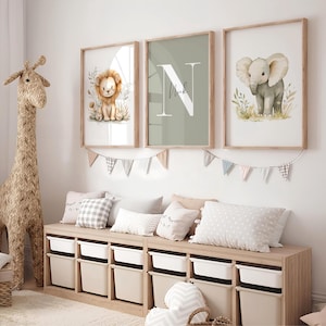 Könnte beinhalten: Drei gerahmte Kunstdrucke für das Kinderzimmer mit einem Löwen, einem Elefanten und dem Buchstaben "N" mit dem Namen "Nick". Die Drucke sind in einem hellen Holzrahmen. Eine gewebte Giraffe und eine Aufbewahrungsbank mit Kissen und Behältern befinden sich im Vordergrund.