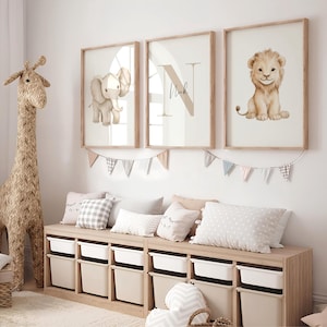 Könnte beinhalten: Drei gerahmte Kunstdrucke für das Kinderzimmer mit einem Elefanten, dem Buchstaben "N" mit dem Namen "Nick" und einem Löwenjungen. Die Drucke sind in einem hellen Beige gehalten und haben Holzrahmen. Darunter befindet sich eine Holz-Aufbewahrungsbank mit weißen Behältern und dekorativen Kissen.