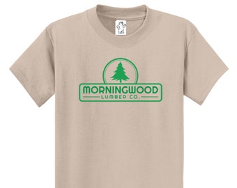 May include: A tan t-shirt featuring a green logo that reads "MORNINGWOOD LUMBER CO." with a green pine tree graphic. The shirt has short sleeves and a crew neck. A small white tag with a bear graphic is on the upper right.