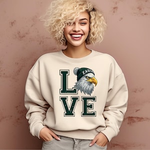 Könnte beinhalten: Cremefarbenes Sweatshirt mit dem Wort "LOVE" in dunkelgrünen Blockbuchstaben. Der Buchstabe "L" befindet sich neben einer Grafik eines Adlers mit einer grünen Mütze. Der Adler hat weiße Federn und einen gelben Schnabel.