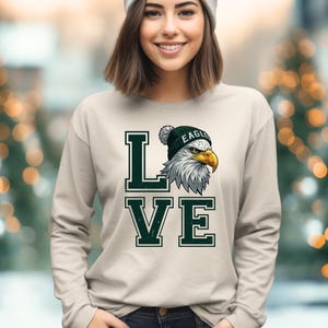 May include: A light beige long-sleeved top with the word "LOVE" in dark green block letters. The letter "L" features an illustration of an eagle wearing a green beanie with the word "EAGLE" on it.