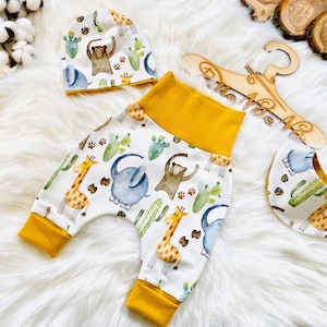 Puede incluir: Un conjunto de ropa para bebé con un estampado de animales de la selva sobre un fondo blanco y amarillo. El conjunto incluye un gorro, pantalones y un babero. Los animales que aparecen son elefantes, jirafas y osos.