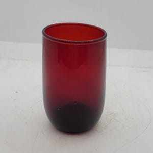 Puede incluir: Un vaso cilíndrico de vidrio rojo rubí con base redondeada. El vidrio hace la transición de un rojo oscuro y profundo en la parte inferior a un rojo más claro y transparente en la parte superior. El vaso está vacío y sobre una superficie blanca.