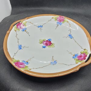 Puede incluir: Una fuente de cerámica blanca con borde y asas doradas. La fuente está decorada con rosas rosas y flores azules. Una pieza decorativa para servir alimentos.