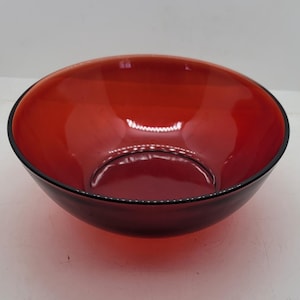 May include: A round, red glass bowl with a black rim. The bowl is empty and sits on a white surface. The bowl's interior reflects light, highlighting its smooth surface. This bowl is suitable for serving food.