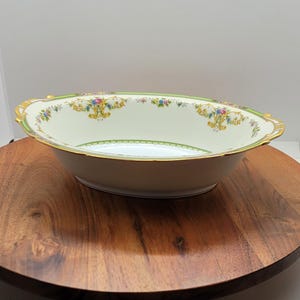 Może przedstawiać: Owalna miska do serwowania w kolorze złamanej bieli z kwiatowym wzorem i złotym wykończeniem. Miska ma zieloną obwódkę i jest prezentowana na drewnianym, okrągłym stojaku. Wzór kwiatowy zawiera żółte, różowe i niebieskie kwiaty.