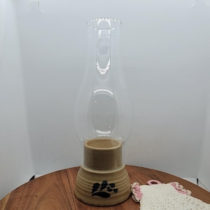 May include: A clear glass oil lamp chimney sits atop a tan ceramic base with a paw print design. A small, white and pink crocheted coaster rests on a wooden surface.