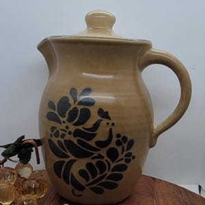 Puede incluir: Jarra de cerámica color canela con tapa y diseño floral negro. La jarra tiene un asa curva y un pico para verter. Una pieza decorativa para la cocina o el comedor.