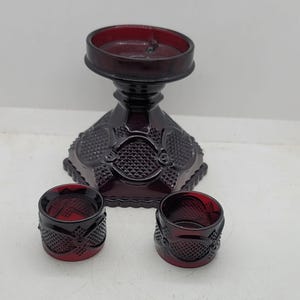 Pode incluir: Um castiçal de vidro vermelho escuro com base de pedestal e dois porta-guardanapos a condizer. O castiçal tem um topo redondo e um padrão decorativo. Os porta-guardanapos são cilíndricos com um design semelhante.