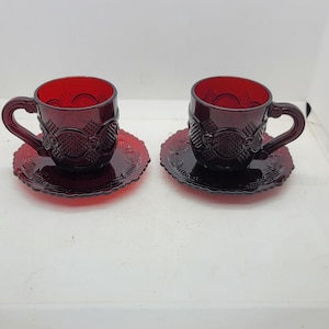 Puede incluir: Dos tazas de té y platillos de vidrio rojo rubí. Las tazas y los platillos tienen un patrón en relieve detallado. Las tazas tienen un asa curva. Los platillos son redondos con un borde festoneado.