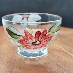 May include: A clear glass bowl with a pedestal base, hand-painted with red, white, and green floral designs. The bowl's rim is slightly flared, and the flowers are arranged around the exterior. The bowl is sitting on a wooden surface.