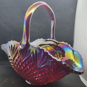 May include: A ruby-red glass basket with a handle, featuring a diamond-cut pattern and iridescent sheen. The rim has a scalloped edge, and the handle arches gracefully. The glass reflects a rainbow of colors, adding to its decorative appeal.