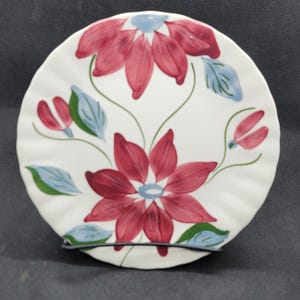 May include: A white ceramic plate with a floral design. The plate features a central red flower with blue accents and green stems and leaves. The plate has a scalloped edge.