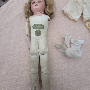 May include: An antique Florodora doll with blonde hair, a porcelain head, and a cloth body featuring a green label that reads Florodora Real Hair Eyebrows. The doll includes a pair of white lace-trimmed bloomers and a pair of white socks.