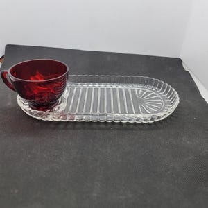 Puede incluir: Una taza de té de vidrio rojo rubí con asa se encuentra sobre una bandeja de servicio de vidrio transparente. La bandeja es de forma ovalada con un diseño acanalado y una sección para la taza. El conjunto es probablemente vintage.