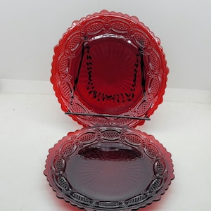 May include: Two red glass serving dishes. The top dish is round, and the bottom dish is oval. Both dishes have a decorative, scalloped edge. The dishes are displayed on a black metal stand against a white background.