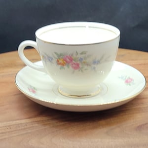 May include: A white teacup and saucer with a floral pattern in shades of pink, yellow, and blue. The cup and saucer have a gold trim.