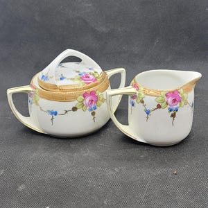 Puede incluir: Un azucarero vintage con tapa y una jarrita de porcelana. Ambas piezas son blancas con ribetes dorados y decoradas con motivos florales rosas y azules. El azucarero tiene un asa.