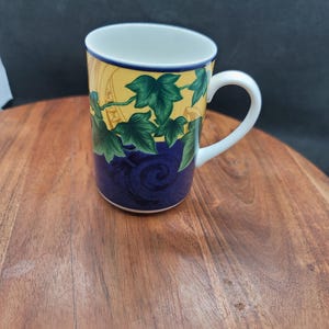 May include: A ceramic mug featuring a two-tone design with a yellow upper section decorated with green ivy leaves and a deep blue lower section with a dark swirling pattern. The mug has a white interior and a white handle on the right side.