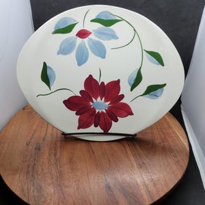 Op de afbeelding: Witte keramische plaat met een bloemenontwerp. De plaat heeft twee blauwe bloemen en één rode bloem met groene bladeren en stelen. De plaat wordt weergegeven op een houten oppervlak.