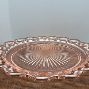 Puede incluir: Una bandeja de servir de vidrio rosa con borde festoneado y diseño de estrella en el centro. La bandeja redonda está sobre una superficie de madera. Esta bandeja de estilo vintage es ideal para servir aperitivos o postres. Diámetro: unos 25 cm.