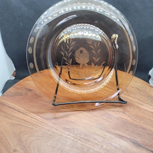 May include: A clear glass plate with a decorative etched floral design in the center. The plate has a gold rim with small gold dots. It is displayed on a black metal stand, set against a dark background.