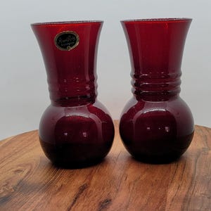 Può includere: Due vasi in vetro rosso rubino con base bulbosa e parte superiore svasata. I vasi sono esposti su una superficie di legno. Un vaso ha un'etichetta dorata con la scritta "Royal Ruby".