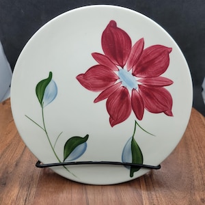 May include: A white ceramic plate with a hand-painted red flower design. The flower has a light blue center and is surrounded by green leaves and blue accents. The plate is displayed on a black metal stand.