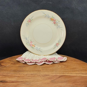 May include: A white ceramic plate with a gold rim and a floral design featuring pink, yellow, and blue flowers. The plate is sitting on a wooden surface.
