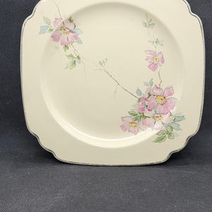 Op de afbeelding: Een wit keramiek bord met een gegolfde rand en een bloemenontwerp met roze en blauwe bloemen. Het bord heeft een zilveren rand.