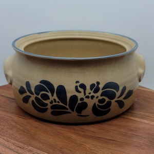 May include: Tan ceramic serving bowl with a blue rim and handles. The bowl features a dark blue floral pattern around the exterior. The bowl is sitting on a wooden surface.